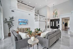 A spacious living room with a grey sofa set, a round coffee table, and a large flat-screen TV. A staircase with a railing leads to an upper level. - 6br Elegant Hollywood Lakes Mansion W/ Pool & Spa 6 Bedroom Home (Hollywood)