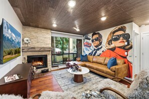 A modern living room with a fireplace, a large TV, and a mural of a couple on a ski lift. - Designer Condo w/ Hot Tub & Ski Locker (Park City)