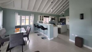 A spacious living area with a dining table and chairs, a kitchen, and a large window with curtains. - The Crest - Serenity Oasis Villa (St. Ann's Bay)