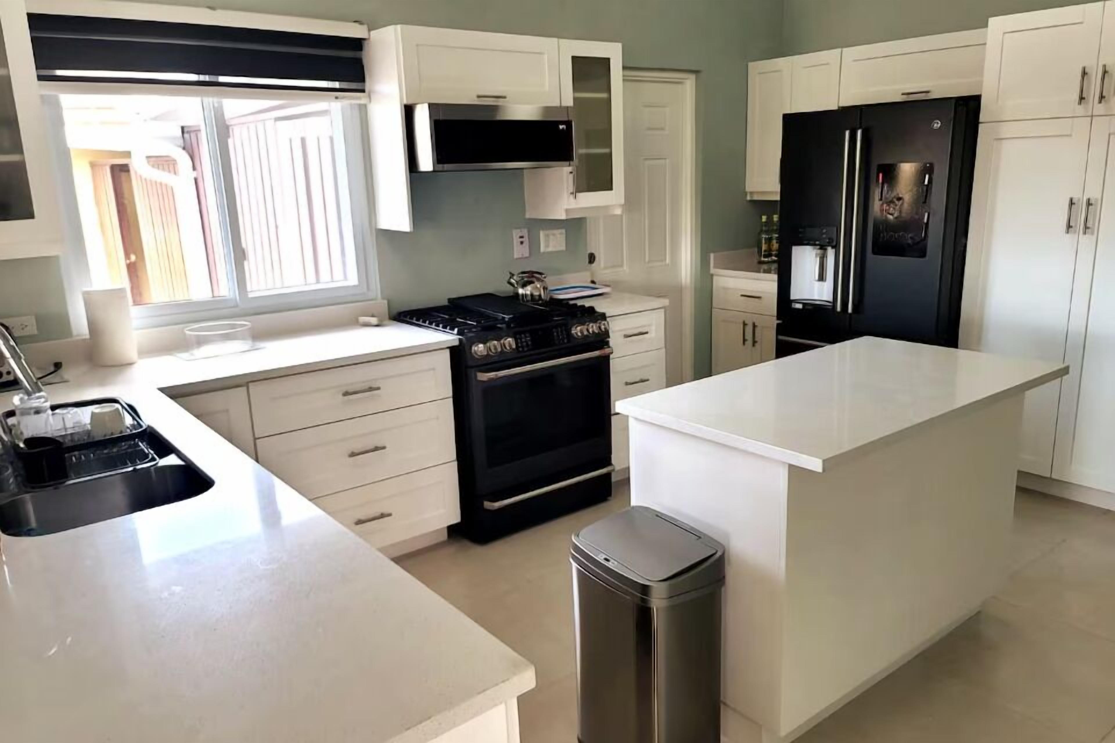 Una cocina con gabinetes blancos, una estufa negra, un bote de basura de acero inoxidable y un refrigerador.