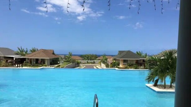 A large swimming pool with a clear view of the ocean in the background, surrounded by resort-style buildings. - The Crest - Serenity Oasis Villa (St. Ann's Bay)