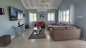 A spacious living room with a high ceiling, a large window, a ceiling fan, and a television mounted on the wall. - The Crest - Serenity Oasis Villa (St. Ann's Bay)