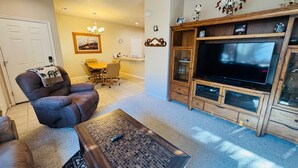 A living room with a leather sofa, a coffee table, a TV on a wooden stand, a dining area with a table and chairs, and a chandelier. - Juniper Retreat | Ground 2BR Pet-Friendly + over $500 in complimentary activities! (Mesquite)