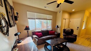 A living room with a brown leather sofa, a coffee table, a desk with a lamp, a ceiling fan, and a window with blinds. - Juniper Retreat | Ground 2BR Pet-Friendly + over $500 in complimentary activities! (Mesquite)