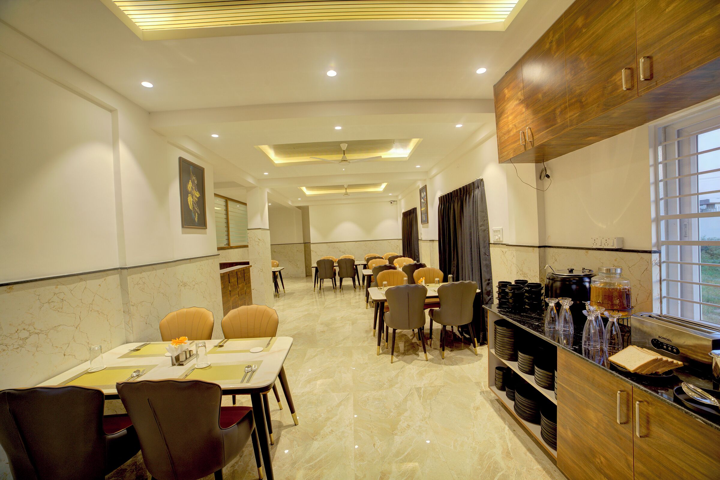 A hotel dining area with multiple tables and chairs, a kitchen counter with appliances, and a view of the outdoors through a window.