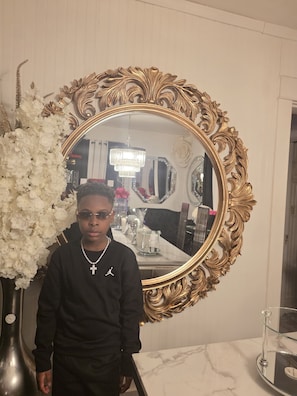 A person standing in front of a large ornate mirror with a gold frame, a white marble countertop, and a vase of white flowers.