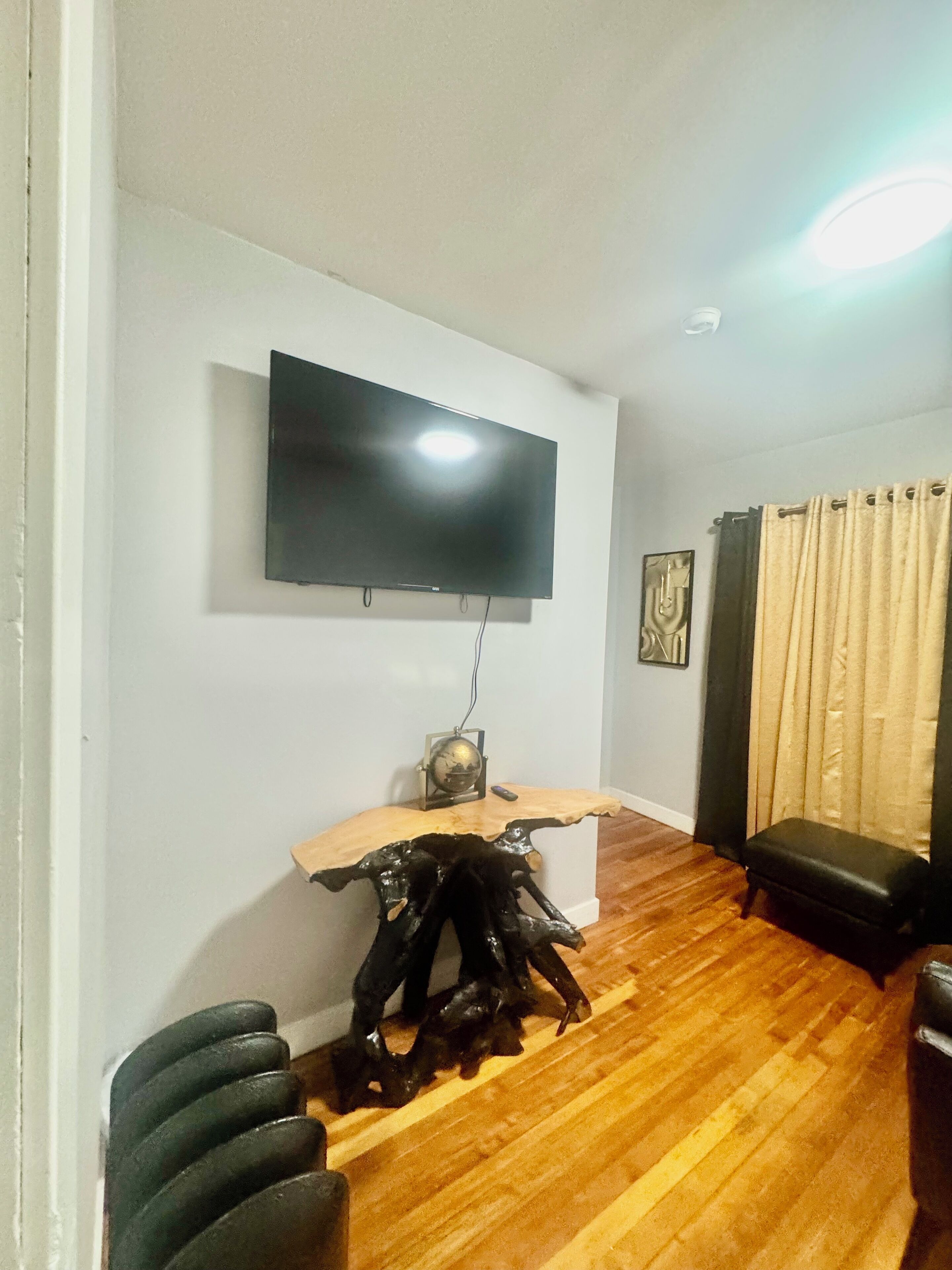 Ein Wohnzimmer mit flachbildfernseher an der Wand, einem Holztisch mit Globus und Fernbedienung, einer schwarzen Bank und einem Vorhang.