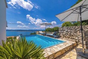 Une piscine avec des chaises longues et des parasols, un mur en pierre et une vue sur la mer.