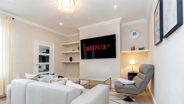 Ett modernt vardagsrum med en platt-TV, en grå fåtölj, en soffa, ett soffbord i glas och en lampa.