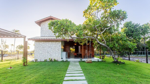 A house with a stone exterior, a paved pathway leading to the entrance, and a well-maintained lawn. - Well-located Condominium in Campinhos in Maraú - RVEs (Maraú)