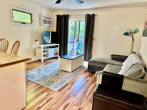 Ein Wohnzimmer mit Holzboden, einer Couch, einem Fernseher, einem Schreibtisch und einem Fenster mit Vorhängen.