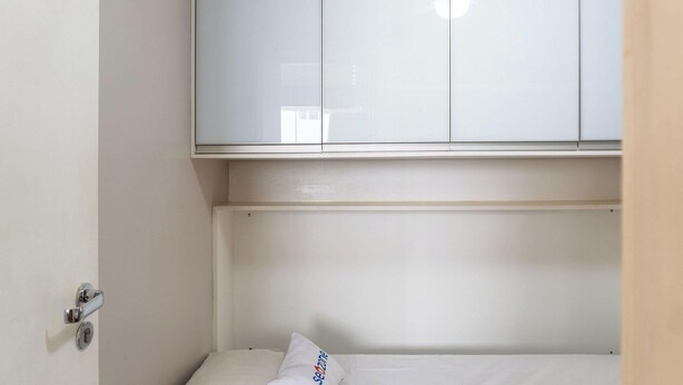A small, neatly made bed with a white mattress and a single pillow, a white wardrobe with a mirror, and a door with a silver handle. - 2brs apt 950 m fr/ Flamboyant Mall Unq1104 (Goiânia)