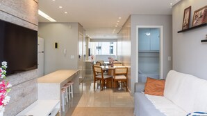 A modern living room with a flat-screen TV, a sofa, a dining area with chairs, and a kitchen in the background. - 2brs apt 950 m fr/ Flamboyant Mall Unq1104 (Goiânia)