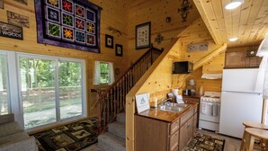 L’intérieur chaleureux d’un chalet avec un escalier en bois, une cuisine équipée d’une cuisinière et d’un réfrigérateur, et une grande fenêtre donnant sur un paysage verdoyant.