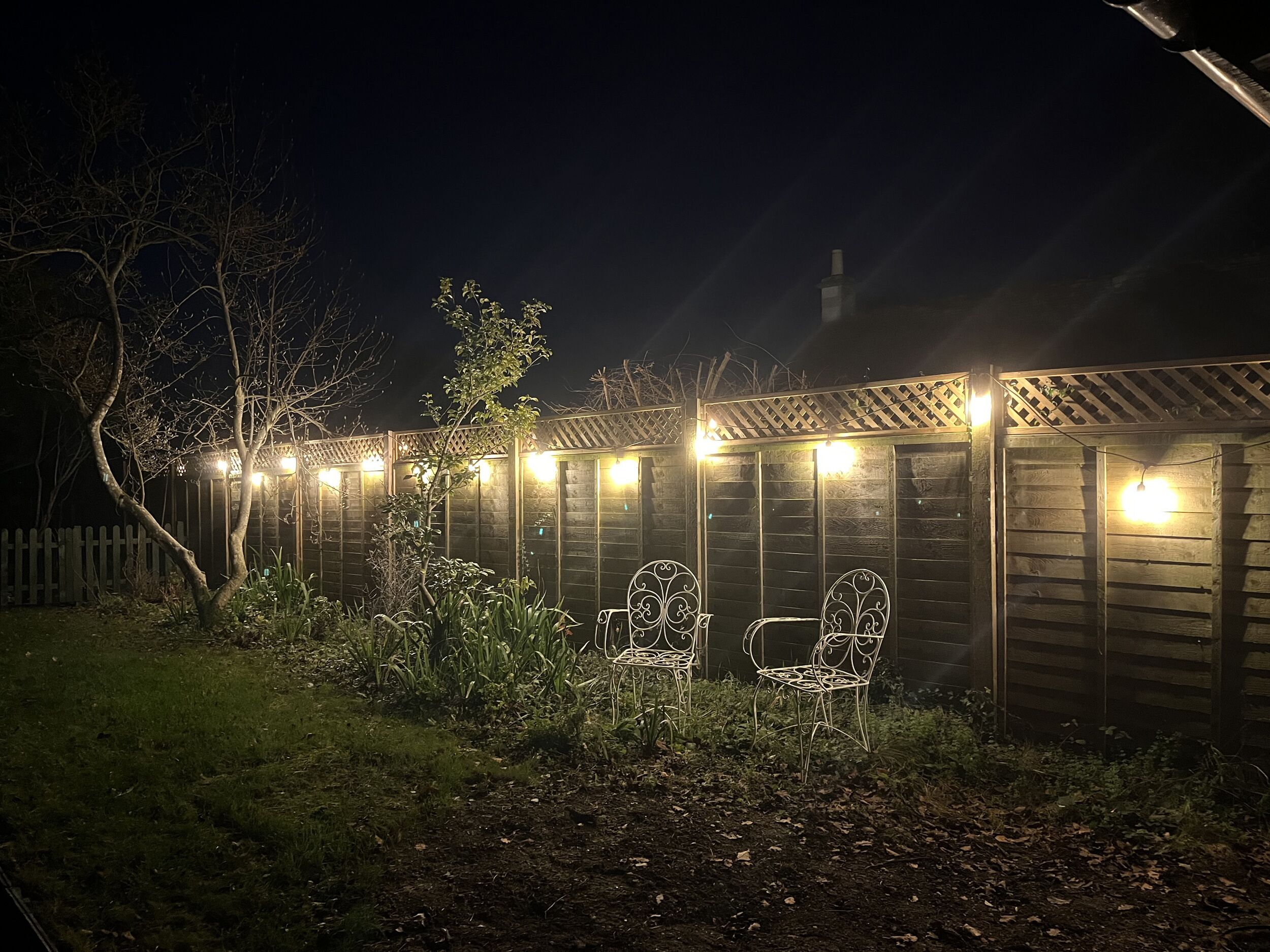 A wooden shed with two metal chairs in front, surrounded by a garden with plants and a white picket fence.