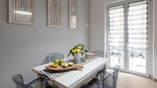 A dining area with a white table, clear glass chairs, a fruit bowl, and striped curtains. - Once in, behind the front door you will find an exclusive atmosphere, finely furnished with shabby chic details and equipped with every comfort.This is La Bonheur!At the entrance a hallway leads to 2 double rooms with beautiful en-suite bathrooms.Perfect (La Spezia)