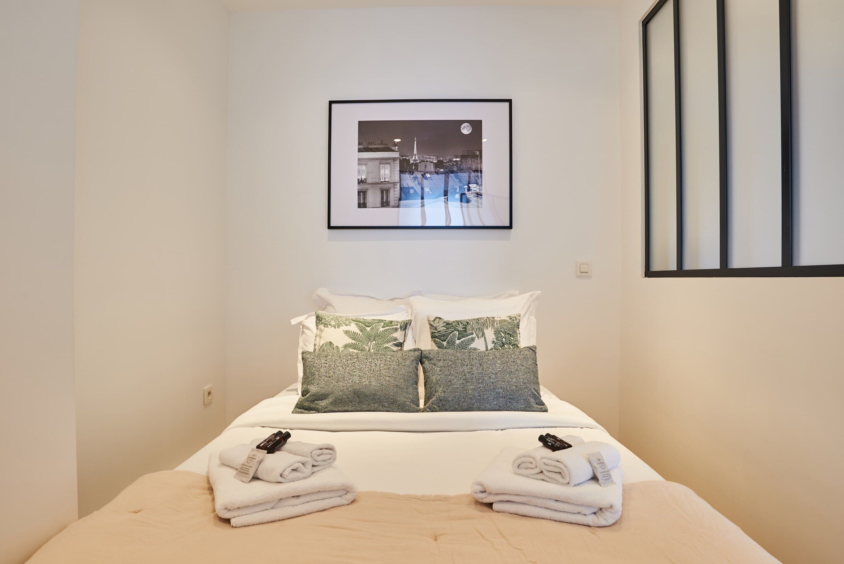 A neatly made bed with white bedding and two pillows, a framed picture of a cityscape on the wall, and a window with blinds.