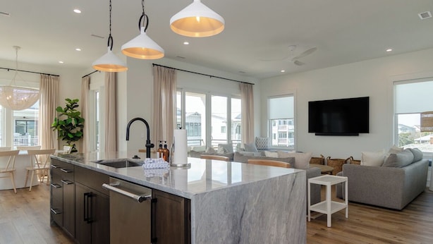 A modern kitchen with a marble island, pendant lighting, and a flat-screen TV mounted on the wall. - Stunning Custom Home with Pool & Rooftop Deck (Wrightsville Beach)