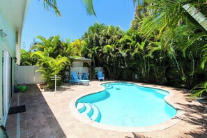 A swimming pool with a curved edge, surrounded by palm trees and a covered seating area with chairs. - Wonderful 3/2 close to beach! Heated pool! (Holmes Beach)