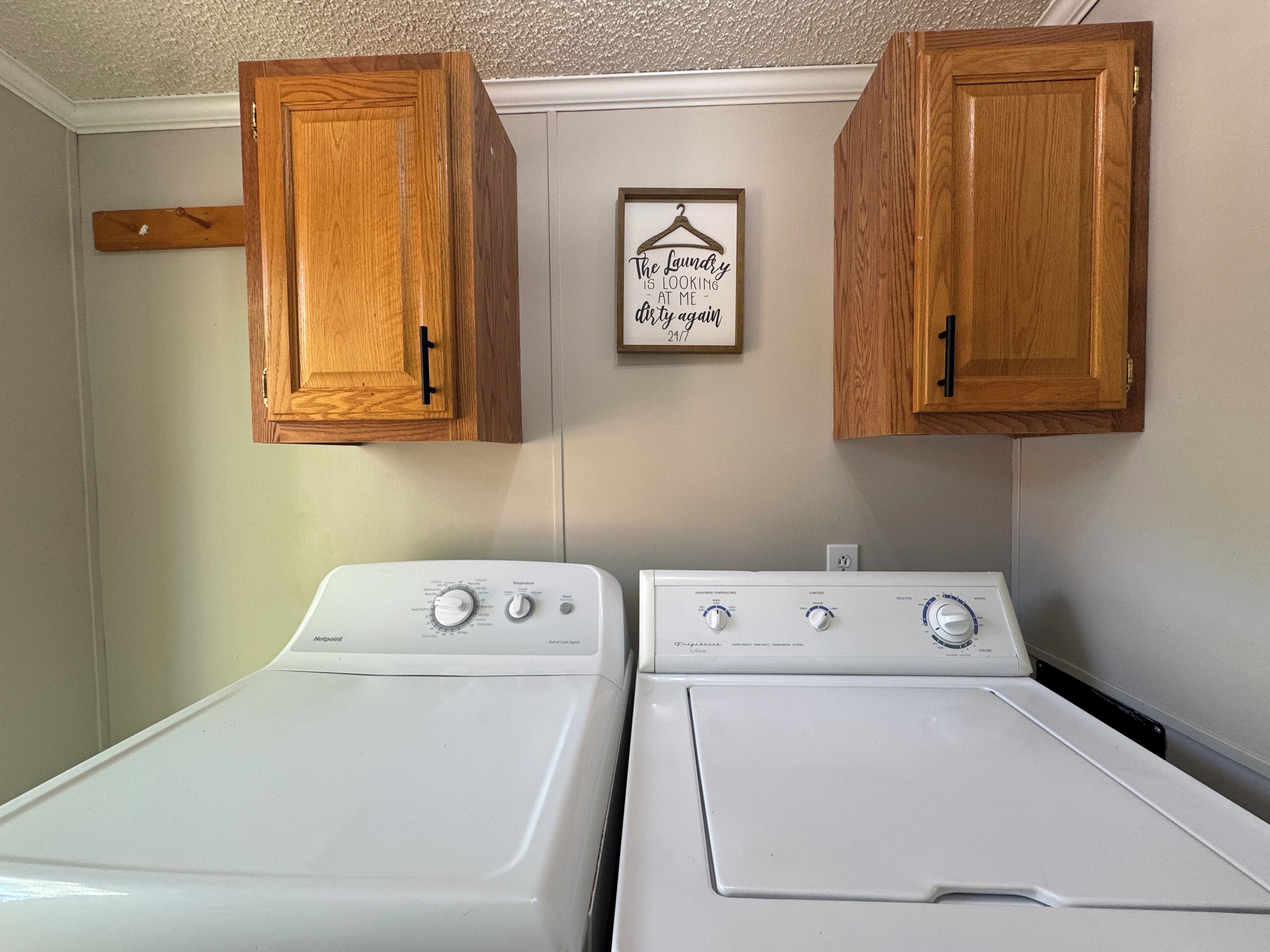 A laundry room with two white front-loading washers and dryers, wooden cabinets above, and a framed quote on the wall.