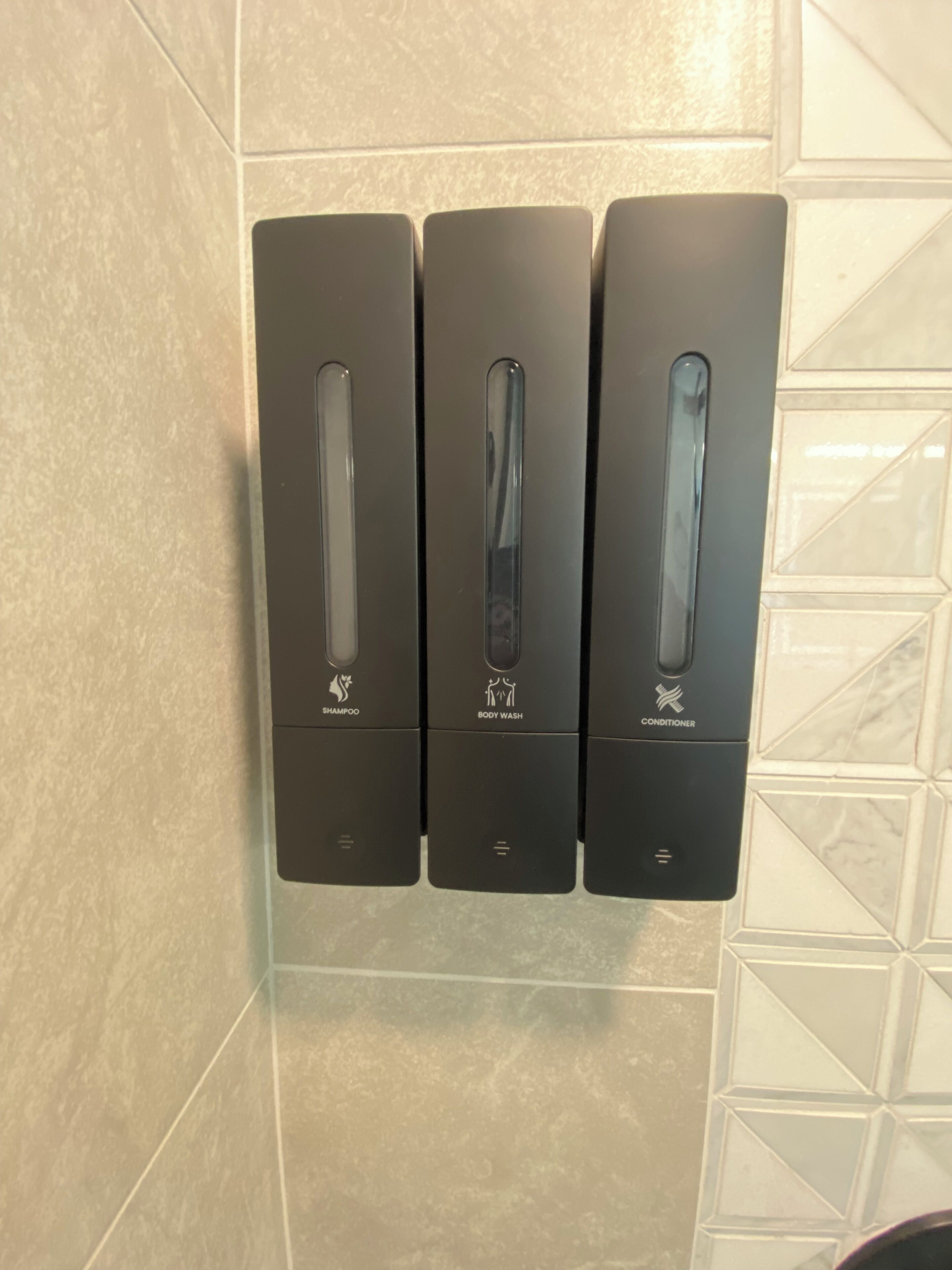 A wall-mounted dispenser with three compartments labeled for shampoo, body wash, and conditioner.