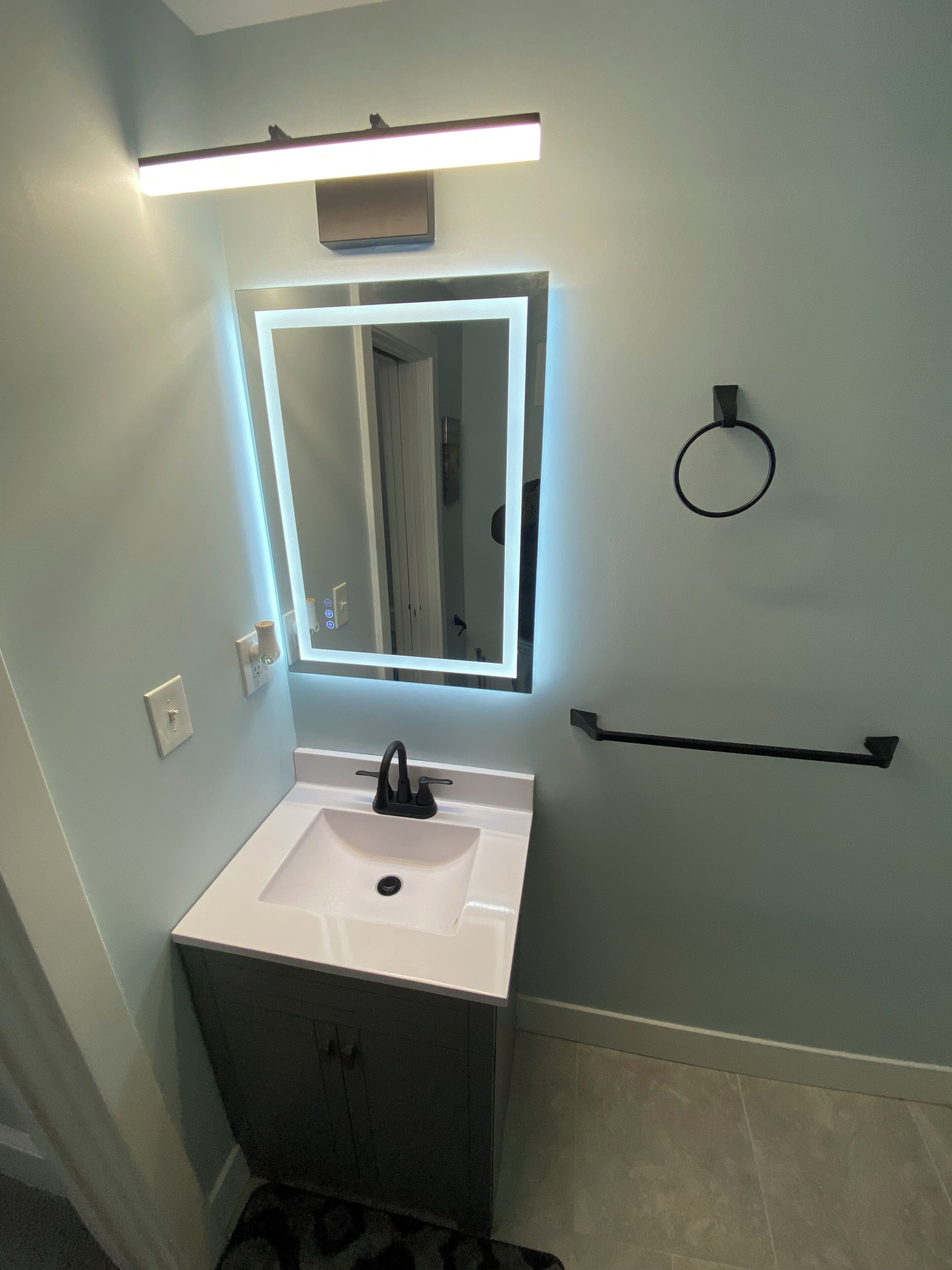 Une salle de bain avec un lavabo blanc, un miroir avec un éclairage au-dessus, un porte-serviettes et un porte-rouleau de papier toilette.