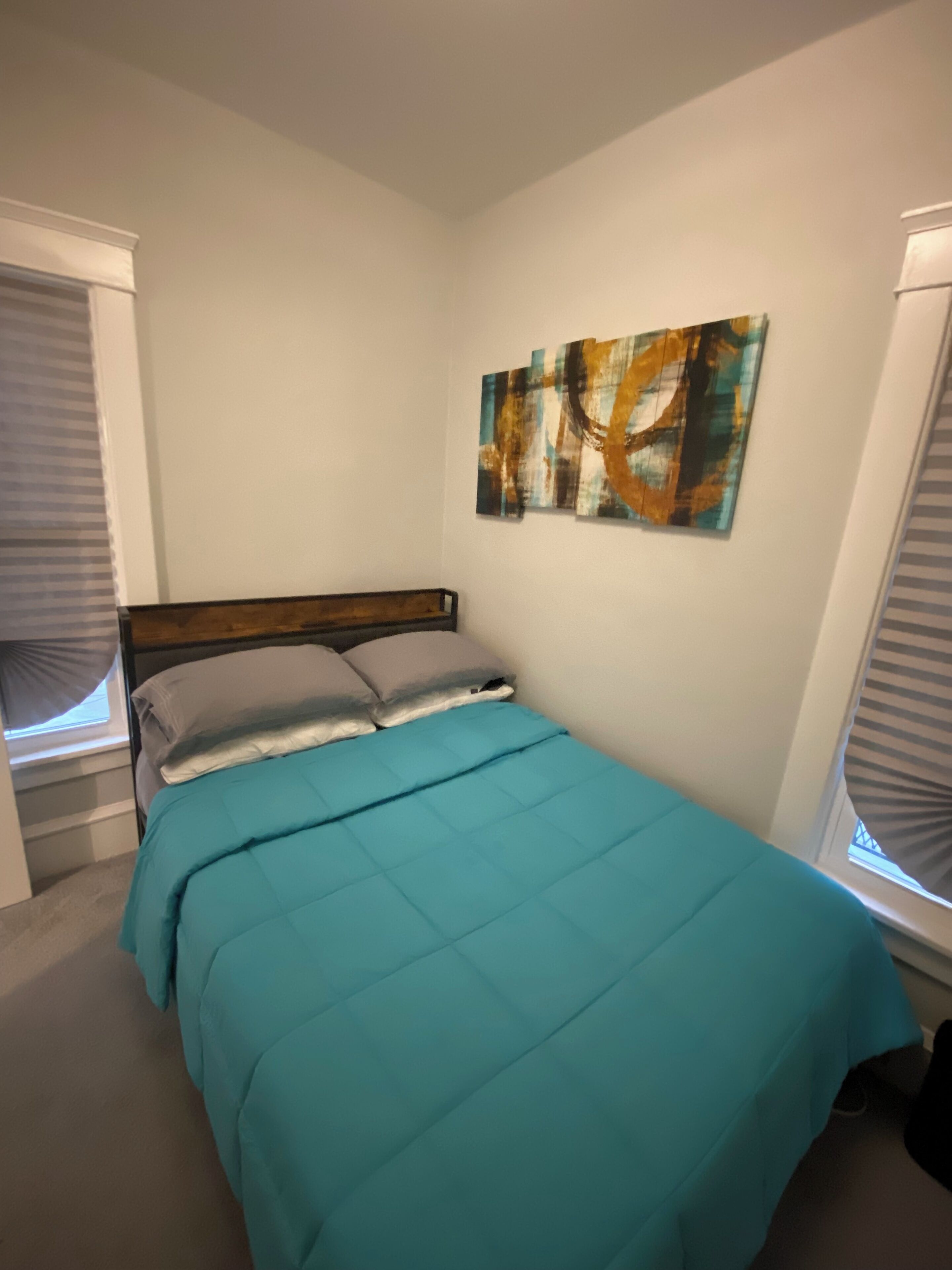 Ein Schlafzimmer mit einem Bett, einem Fenster mit Jalousien und einem an der Wand hängenden abstrakten Gemälde.