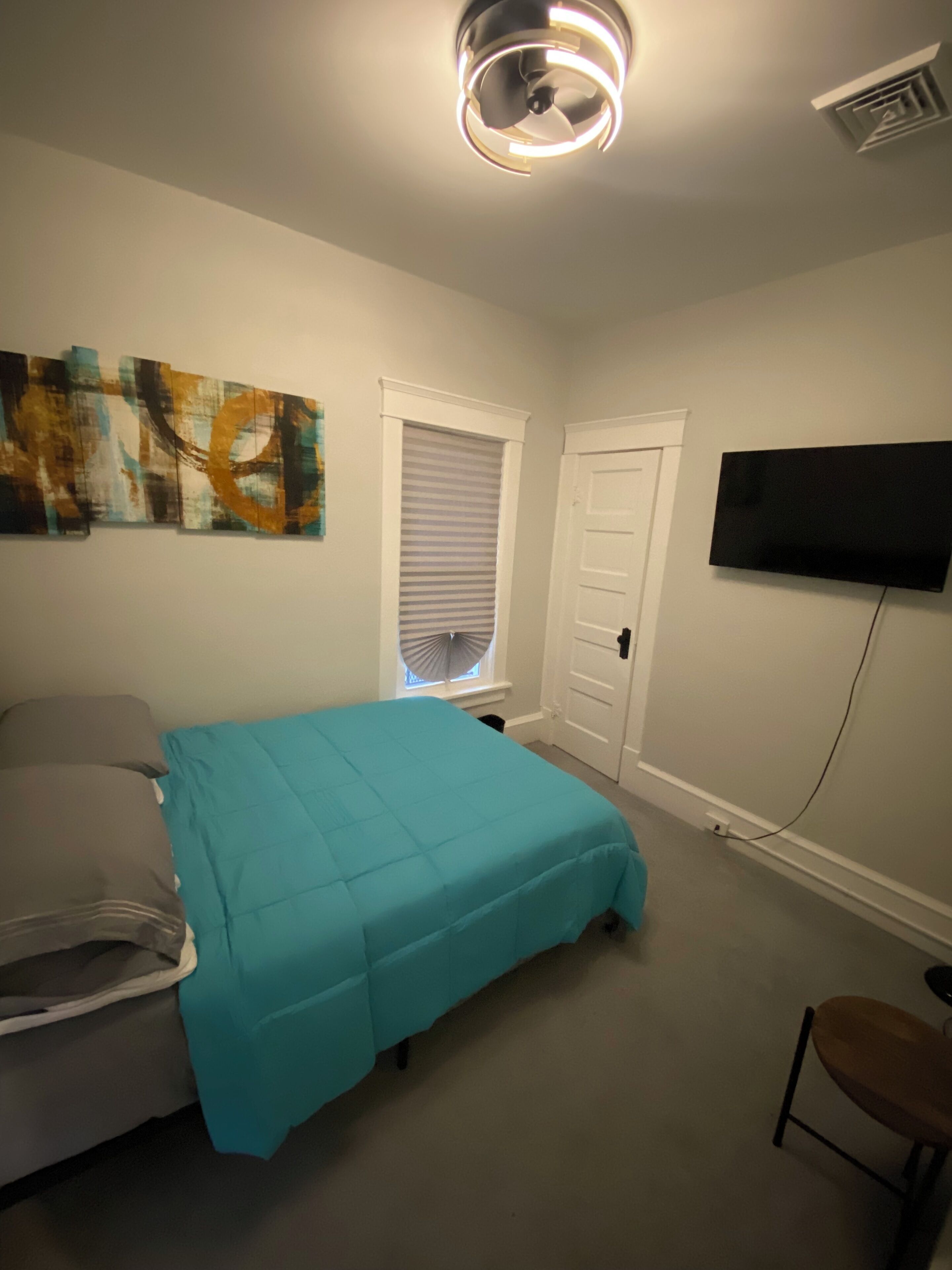 Ein Schlafzimmer mit einem Bett, einem Fenster mit Jalousien, einer Tür, einem Wand-Fernseher und einer Deckenleuchte.