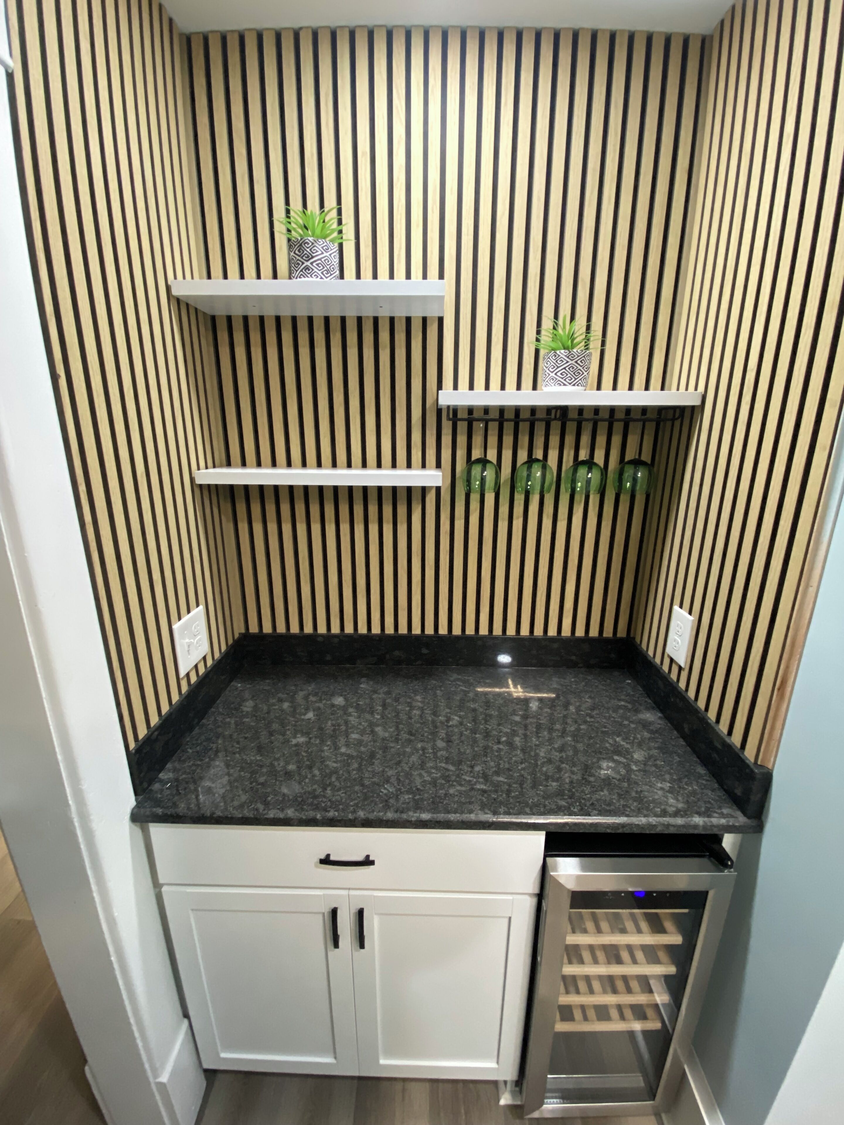 Une cuisine compacte avec un plan de travail noir, des placards blancs et un four encastré. On y trouve deux étagères blanches ornées d’objets décoratifs et une plante en pot.