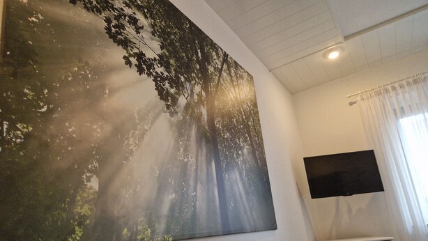 A room with a large wall-mounted artwork depicting a forest scene, a white dresser with drawers, and a flat-screen TV mounted on the wall. - Apartment centrally located to the spa clinic incl. parking lot (Bad Oeynhausen)
