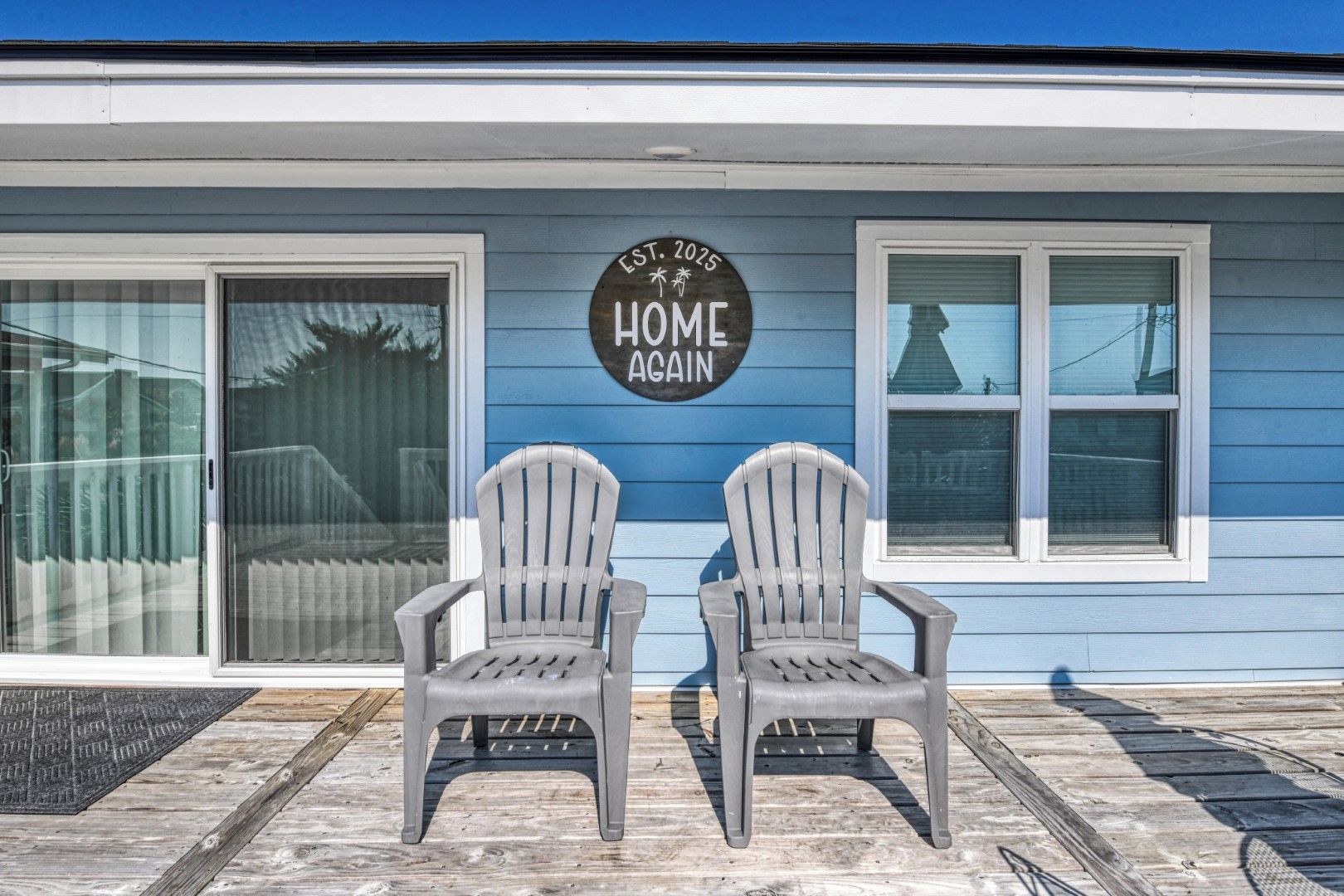 Een huis met een bord waarop staat 'EST. 2025 HOME AGAIN' en twee Adirondack-stoelen ervoor.