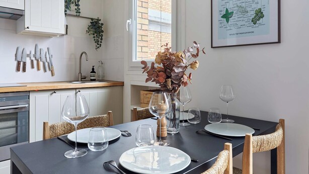 Uma sala de jantar com uma mesa posta para quatro pessoas, um vaso com flores secas e uma ilustração botânica emoldurada na parede.