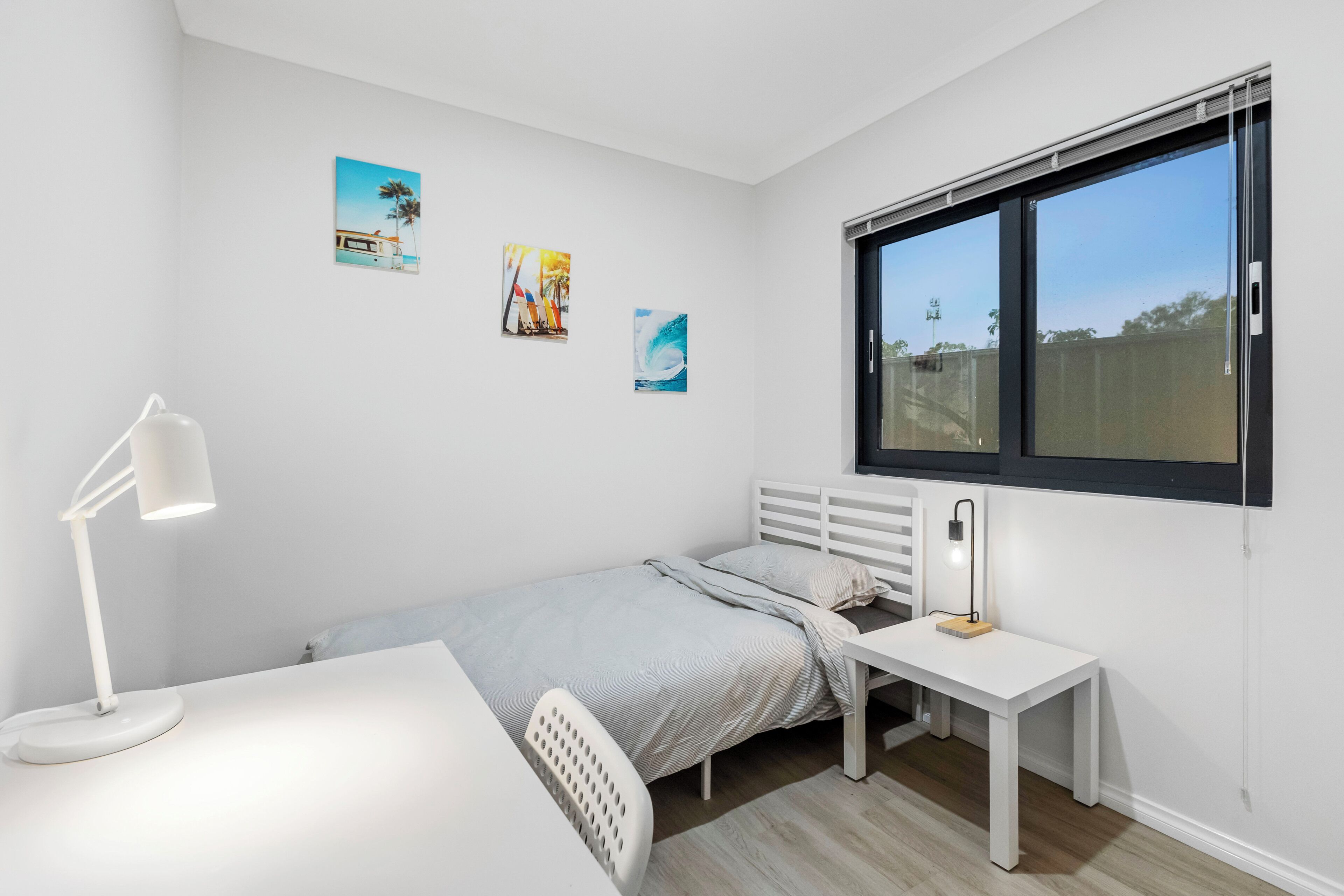 A bedroom with a single bed, a desk, a chair, a standing lamp, and a window with blinds.