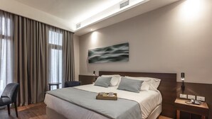 Een moderne hotelkamer met een groot bed, een nachtkastje met een lamp, een stoel en uitzicht op het omliggende landschap door grote ramen.