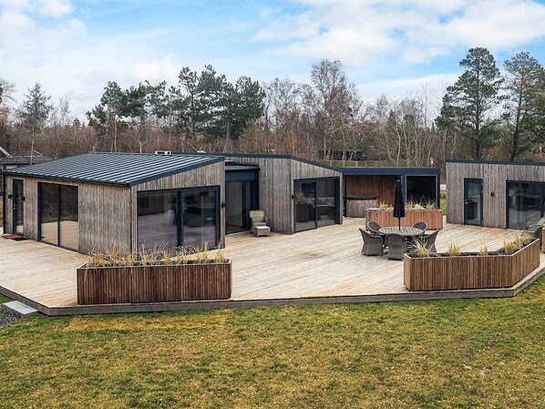 A modern outdoor deck with built-in seating and planters, surrounded by wooden structures with large glass doors. - 5 Star Holiday Home in Vaeggerlose (Vaeggerlose)
