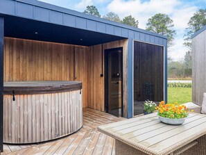 A hot tub with a wooden exterior, a glass door, and a patio with a table and flowers. - 5 Star Holiday Home in Vaeggerlose (Vaeggerlose)