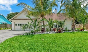 A two-story house with a garage, surrounded by a well-maintained lawn and various tropical plants.