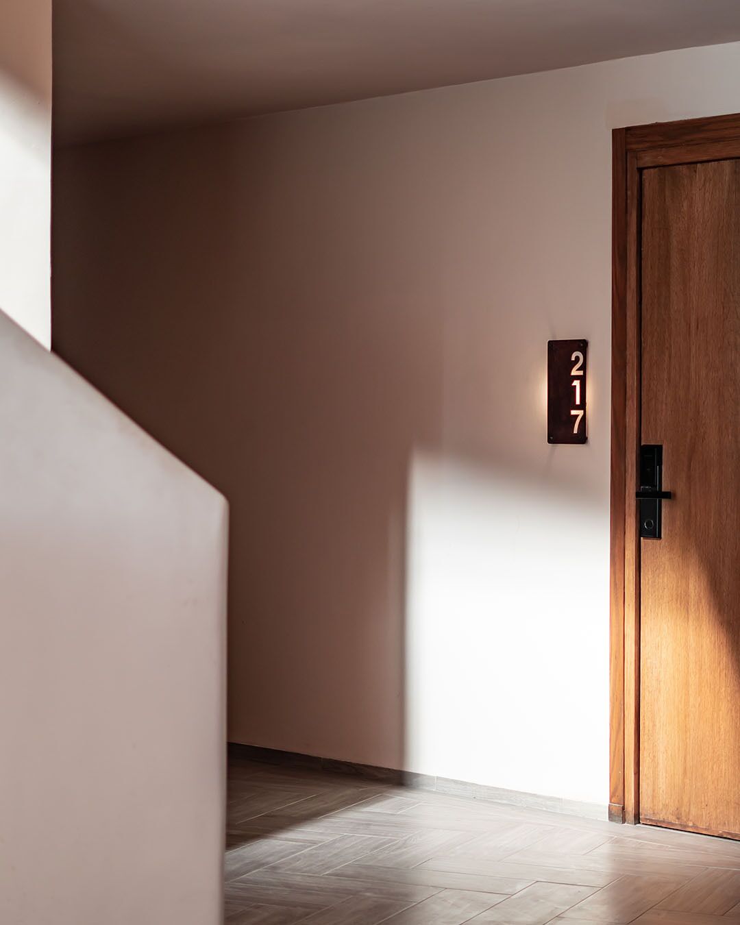 Un pasillo con una puerta de madera marcada con el número 217, un interruptor de luz y un suelo con estampado tipo tablero de ajedrez.