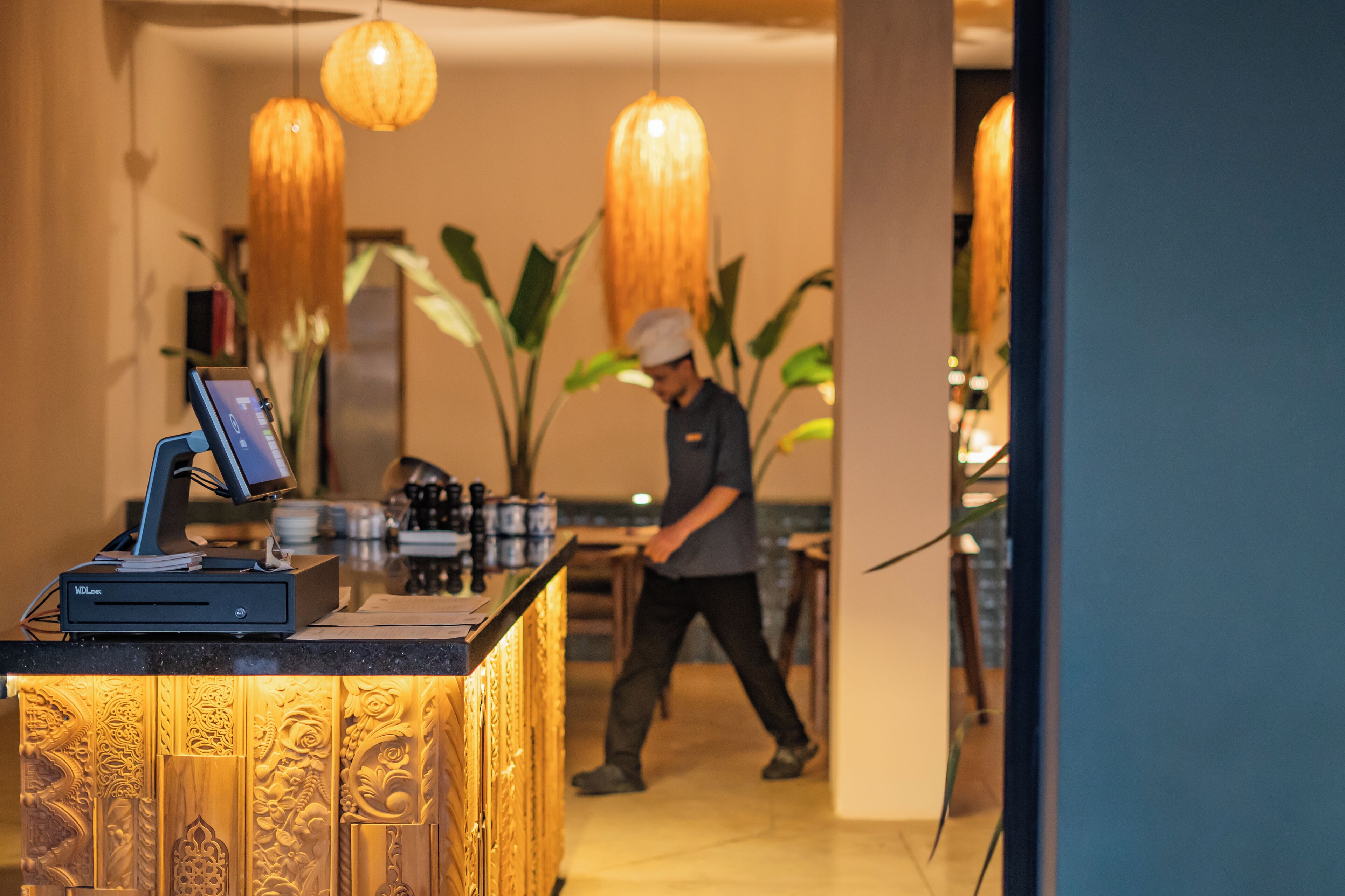 A reception desk with a digital screen, a wooden panel with intricate carvings, and a person walking in the background.