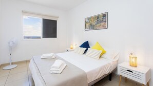 Hotellihuone, jossa on sänky, yöpöydät, lattiatuuletin, ikkuna, jossa on sälekaihtimet, ja seinälle kiinnitetty taideteos.