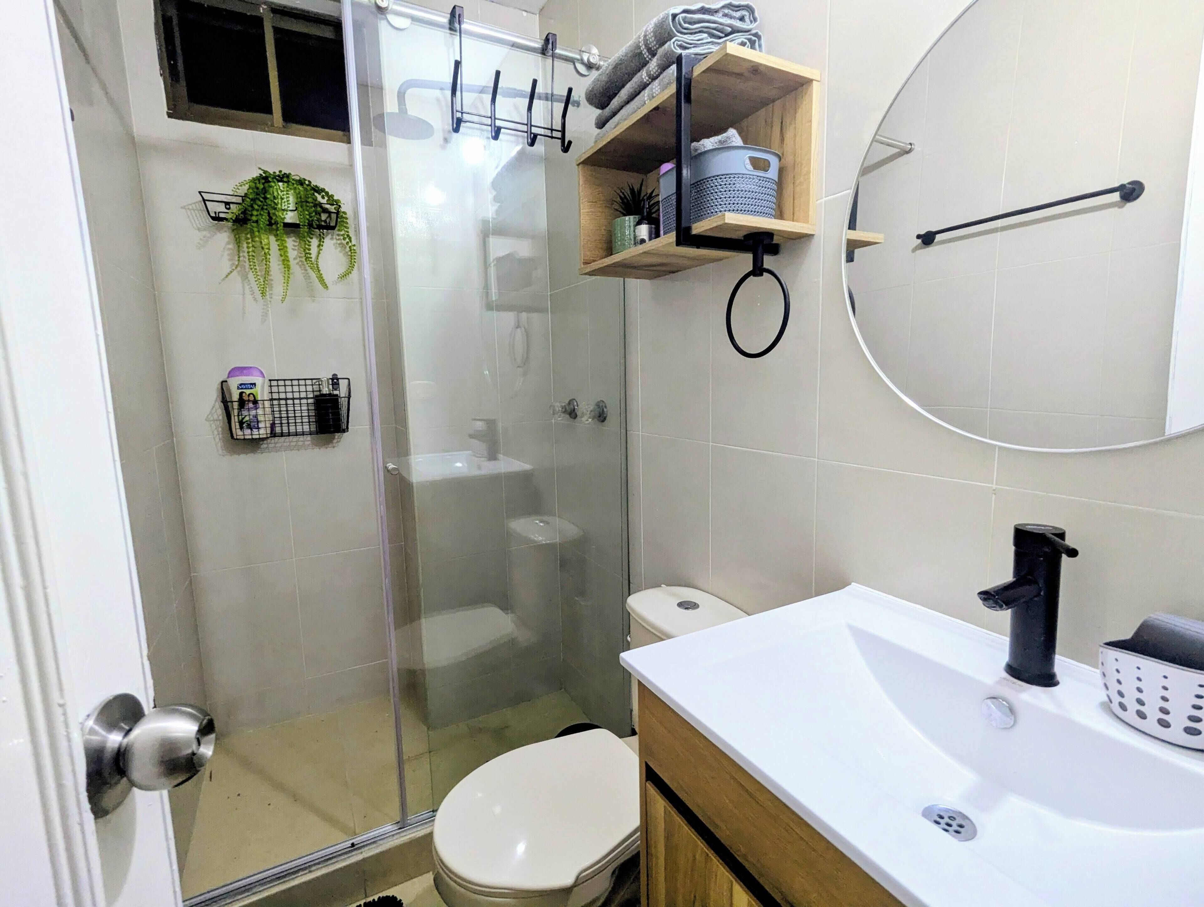 Une salle de bain avec une douche à parois de verre, des toilettes blanches, un lavabo avec un robinet noir et une étagère en bois où sont rangés les articles de toilette.