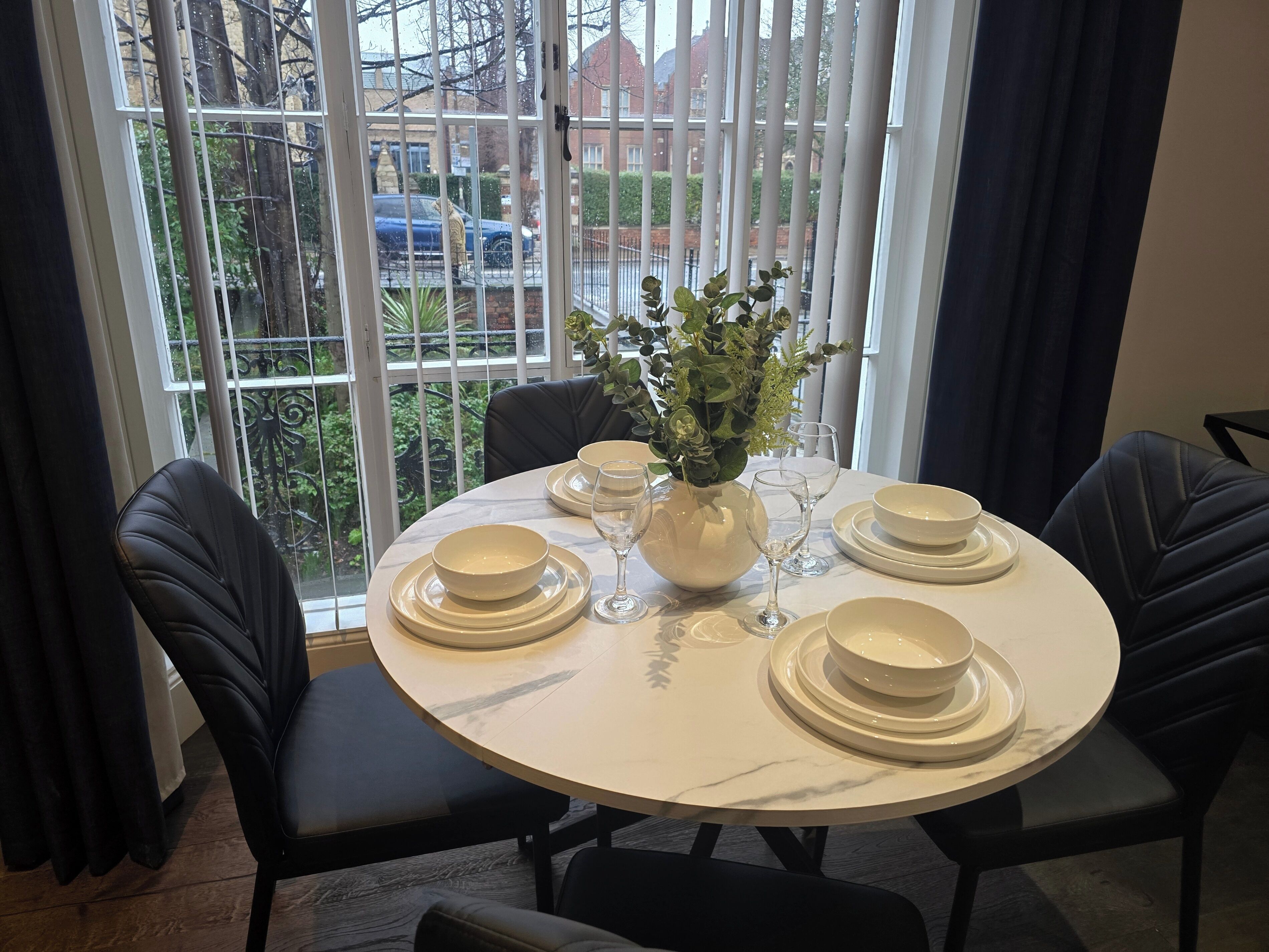 A round dining table with white plates, bowls, and wine glasses, set against a window with a view of a residential area.