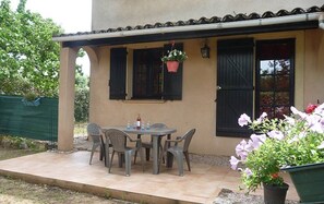 Une terrasse avec une table et des chaises, un pot de fleurs suspendu et un bâtiment avec des volets noirs.