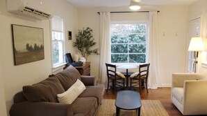 Ein Wohnzimmer mit braunem Sofa, einem kleinen Tisch, einem weißen Sessel, einem Esstisch mit Stühlen und einem großen Fenster mit Vorhängen.