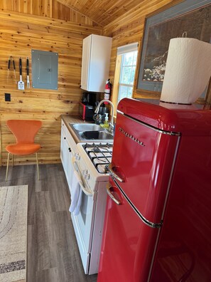 Una cocina compacta con paredes de madera, un refrigerador rojo, una estufa, un fregadero y una pequeña silla naranja.