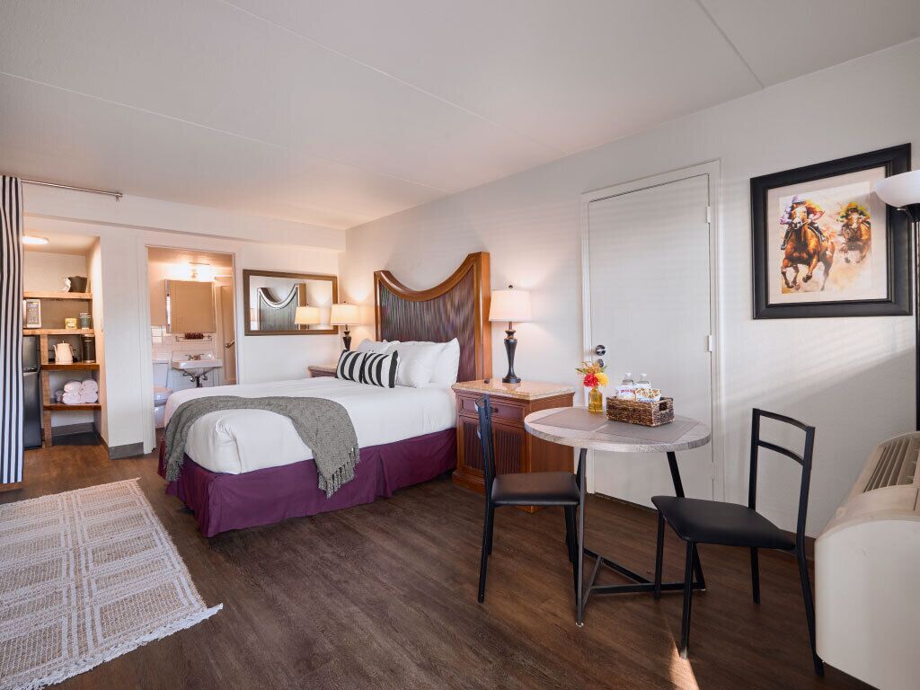 Habitación de hotel con cama, una pequeña mesa redonda con sillas, escritorio, lámpara, espejo y un cuadro en la pared.
