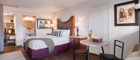 A hotel room with a bed, a small round table with chairs, a desk, a chair, a lamp, a mirror, and a framed picture on the wall.