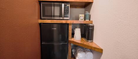 A compact kitchen with a microwave, a refrigerator, a coffee maker, and a kettle on wooden shelves.