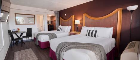 A hotel room with two beds, a desk, chairs, a lamp, and a window.