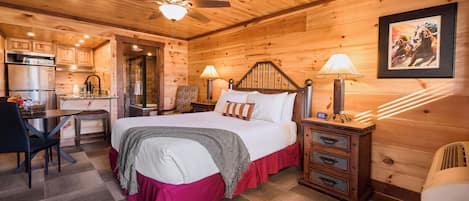 A wooden cabin interior with a bed, bedside lamps, a desk with a chair, and a kitchen area.